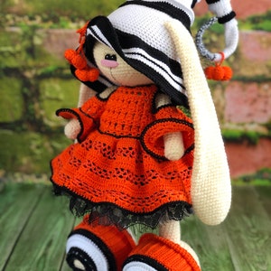 Puede incluir: Un conejito de crochet con un sombrero a rayas blanco y negro, un vestido naranja con ribetes negros y zapatos a rayas blanco y negro.
