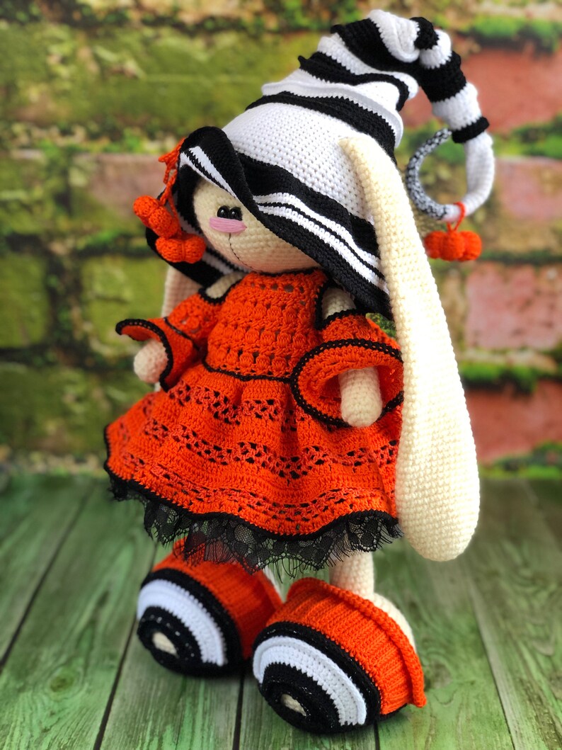 Puede incluir: Un conejito de crochet con un sombrero a rayas blanco y negro, un vestido naranja con ribetes negros y zapatos a rayas blanco y negro.