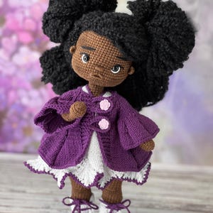 May include: A crocheted doll with brown skin, black curly hair, and a purple jacket. The doll is wearing a white dress with a lace trim and white shoes with purple laces.