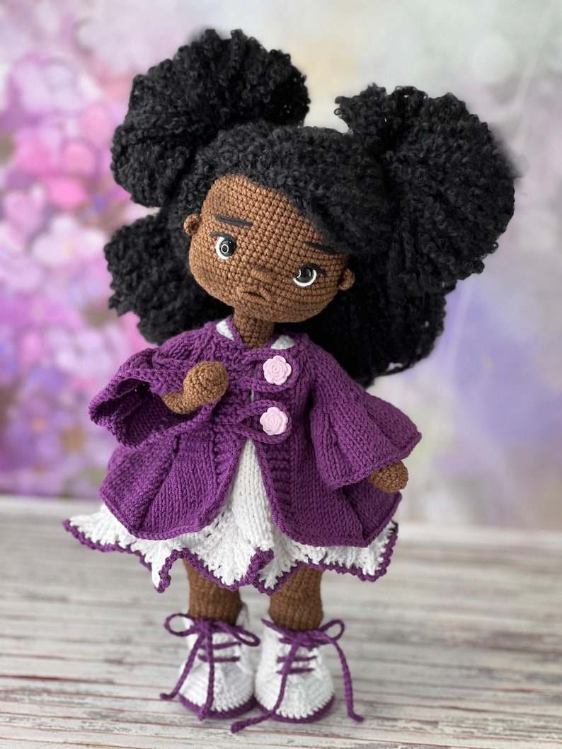 May include: A crocheted doll with brown skin, black curly hair, and a purple jacket. The doll is wearing a white dress with a lace trim and white shoes with purple laces.