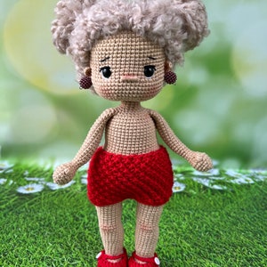 May include: A crocheted doll with blonde curly hair, wearing red shorts and red shoes with white trim. The doll has a neutral expression and is standing on a green grassy surface.