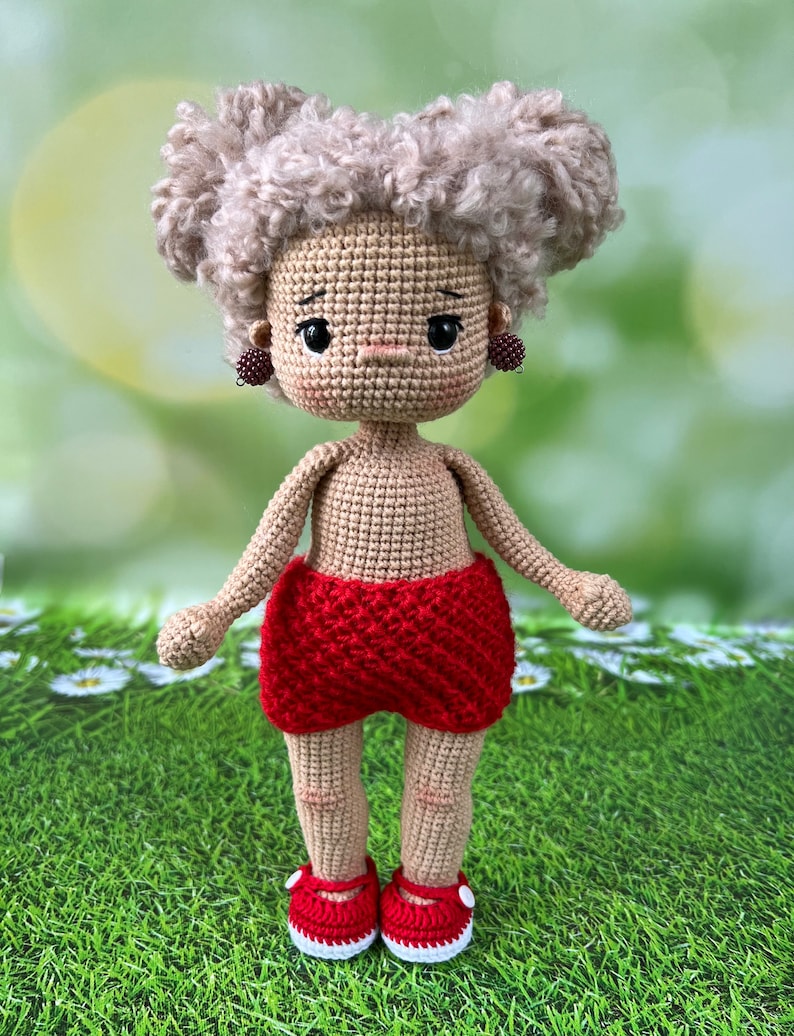 May include: A crocheted doll with blonde curly hair, wearing red shorts and red shoes with white trim. The doll has a neutral expression and is standing on a green grassy surface.
