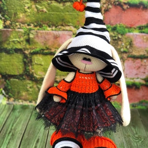 Puede incluir: Un conejito de crochet con un sombrero de bruja a rayas blancas y negras con un peque&ntilde;o pomp&oacute;n naranja, un vestido naranja con ribete de encaje negro y botas a rayas blancas y negras.
