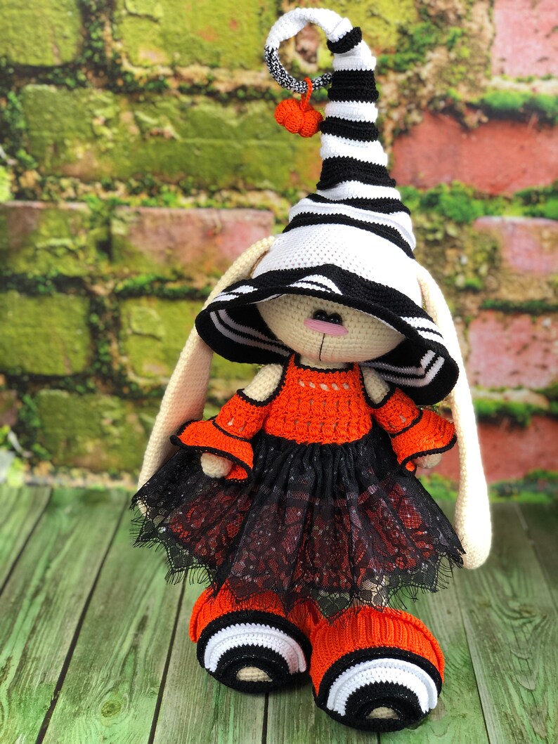 Puede incluir: Un conejito de crochet con un sombrero de bruja a rayas blancas y negras con un peque&ntilde;o pomp&oacute;n naranja, un vestido naranja con ribete de encaje negro y botas a rayas blancas y negras.