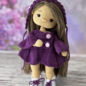 May include: A crocheted doll with long brown hair, wearing a purple hooded jacket, white boots, and a white hat with a purple trim.