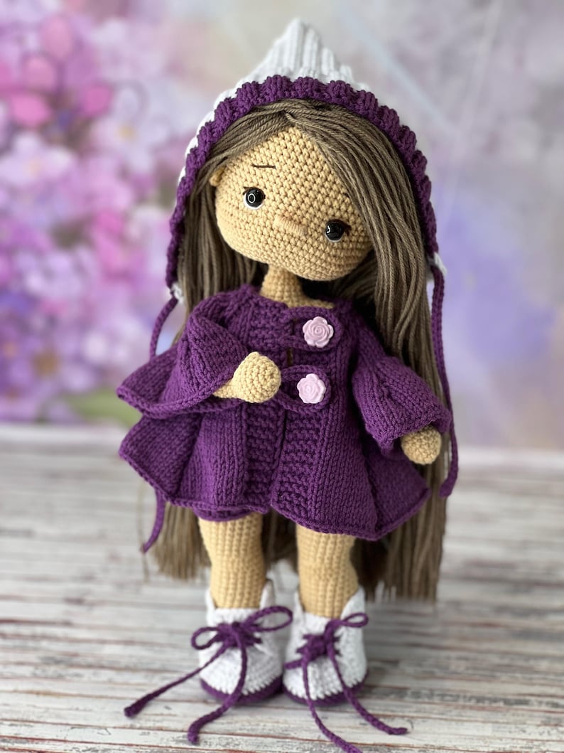 May include: A crocheted doll with long brown hair, wearing a purple hooded jacket, white boots, and a white hat with a purple trim.
