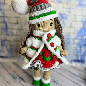 Puede incluir: Una mu&ntilde;eca de crochet que lleva un vestido blanco, rojo y verde, un abrigo blanco y un gorro gris y verde con un pomp&oacute;n blanco. La mu&ntilde;eca tiene el pelo casta&ntilde;o y est&aacute; de pie sobre una superficie de madera blanca.