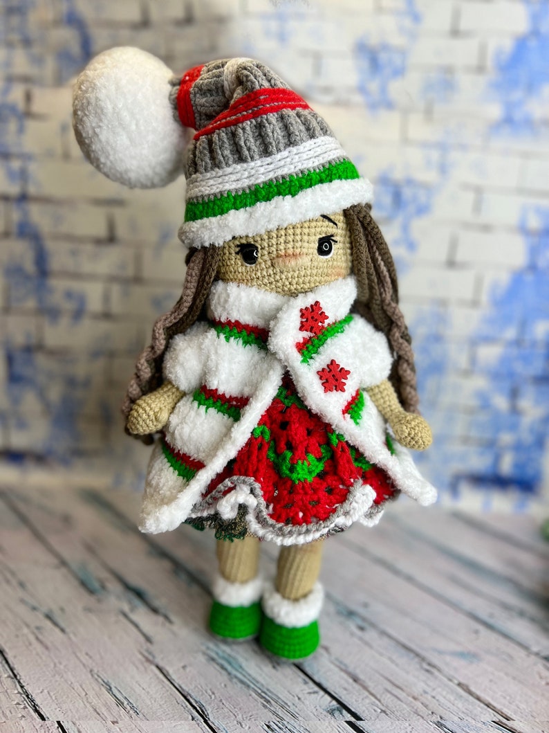 Puede incluir: Una mu&ntilde;eca de crochet que lleva un vestido blanco, rojo y verde, un abrigo blanco y un gorro gris y verde con un pomp&oacute;n blanco. La mu&ntilde;eca tiene el pelo casta&ntilde;o y est&aacute; de pie sobre una superficie de madera blanca.