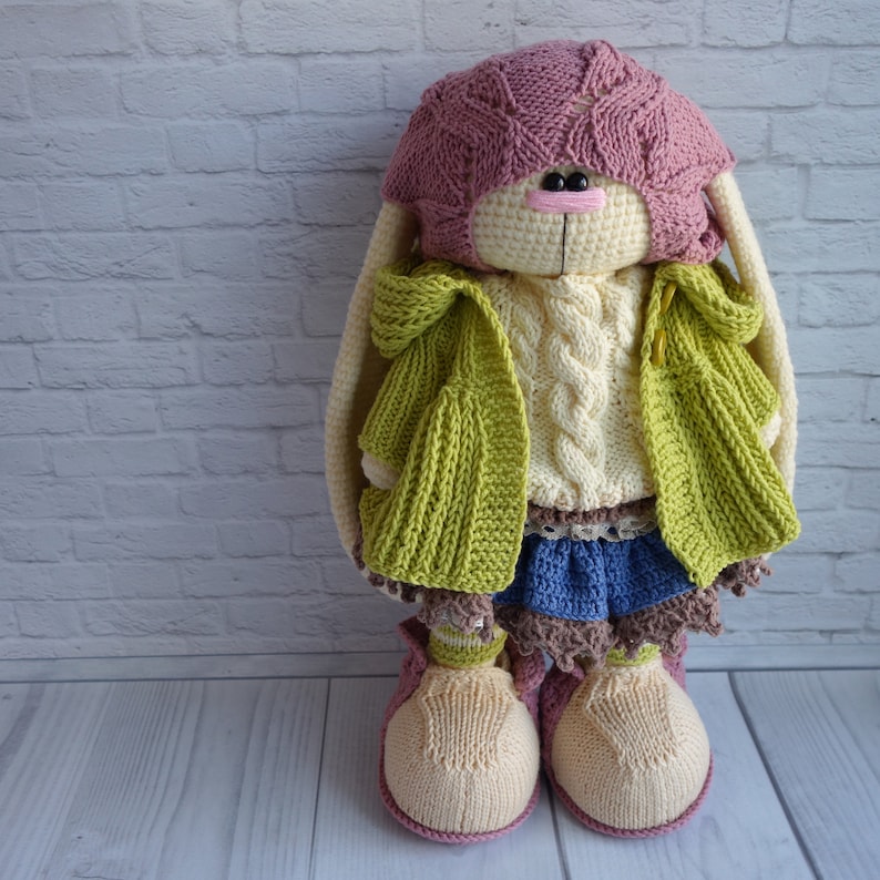 Pode incluir: Um boneco de coelho de croch&ecirc; vestindo um chap&eacute;u rosa, um su&eacute;ter creme, um casaco verde, uma saia azul e sapatos marrons.