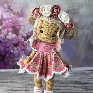 May include: A crocheted doll with blonde hair wearing a pink dress and a flower crown. The doll is standing on a wooden surface.