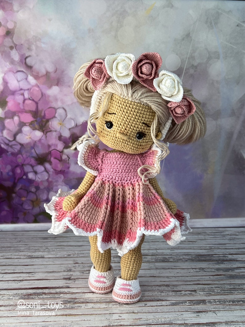 May include: A crocheted doll with blonde hair wearing a pink dress and a flower crown. The doll is standing on a wooden surface.