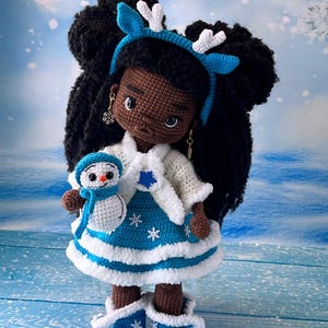 May include: A crocheted doll with brown skin and black hair wearing a blue and white dress with a white fur trim. The doll is holding a small crocheted snowman. The doll is wearing a blue headband with white antlers.