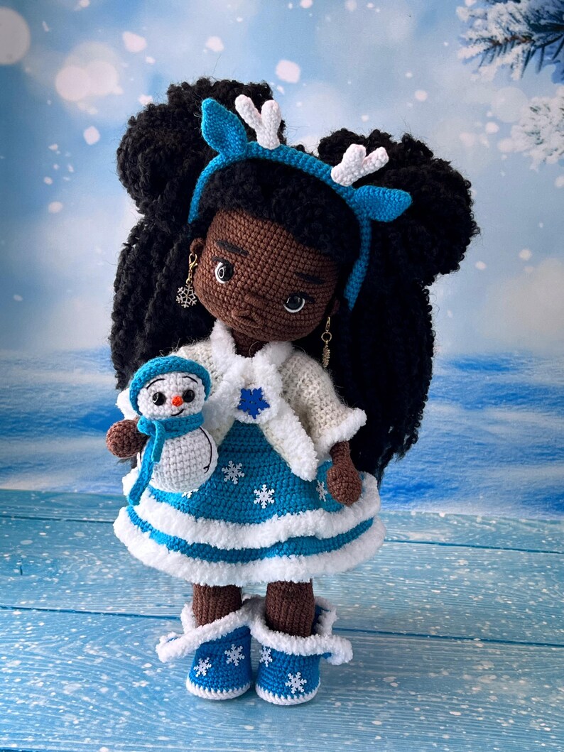 May include: A crocheted doll with brown skin and black hair wearing a blue and white dress with a white fur trim. The doll is holding a small crocheted snowman. The doll is wearing a blue headband with white antlers.