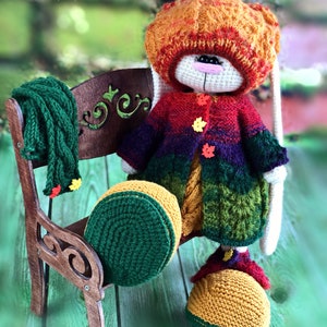 K&ouml;nnte beinhalten: Ein geh&auml;keltes H&auml;schen, das einen Regenbogenpullover, eine gr&uuml;ne M&uuml;tze und gelbe Schuhe tr&auml;gt. Das H&auml;schen sitzt auf einer Holzbank mit einem gr&uuml;nen Schal.