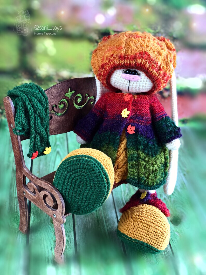 K&ouml;nnte beinhalten: Ein geh&auml;keltes H&auml;schen, das einen Regenbogenpullover, eine gr&uuml;ne M&uuml;tze und gelbe Schuhe tr&auml;gt. Das H&auml;schen sitzt auf einer Holzbank mit einem gr&uuml;nen Schal.