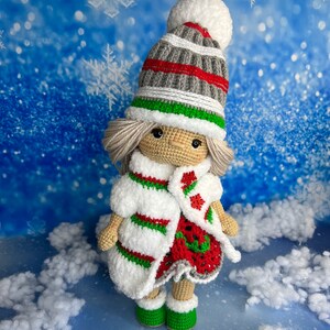 Puede incluir: Una mu&ntilde;eca de crochet con un gorro a rayas blanco, rojo y verde, un abrigo a rayas blanco y rojo y zapatos verdes. La mu&ntilde;eca est&aacute; de pie sobre una superficie blanca y esponjosa sobre un fondo azul con copos de nieve blancos.