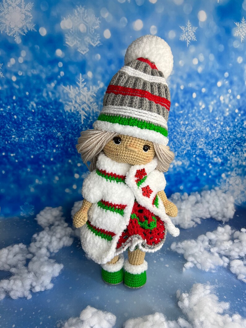 Puede incluir: Una mu&ntilde;eca de crochet con un gorro a rayas blanco, rojo y verde, un abrigo a rayas blanco y rojo y zapatos verdes. La mu&ntilde;eca est&aacute; de pie sobre una superficie blanca y esponjosa sobre un fondo azul con copos de nieve blancos.