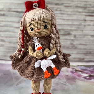 May include: A crocheted doll with blonde hair, wearing a brown dress and red shoes, holding a white crocheted goose.