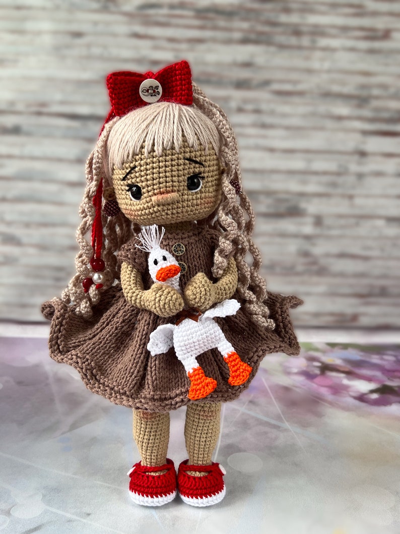 May include: A crocheted doll with blonde hair, wearing a brown dress and red shoes, holding a white crocheted goose.