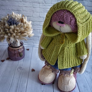 Pode incluir: Um coelho de croch&ecirc; vestindo uma jaqueta verde com capuz e bot&otilde;es amarelos. O coelho tem um rosto rosa e orelhas compridas. O coelho est&aacute; em p&eacute; sobre uma superf&iacute;cie de madeira branca.