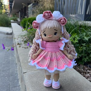 May include: A crocheted doll with pink and white clothing, a pink and white flower crown, and brown hair. The doll is wearing white sneakers and is standing on a gray surface.