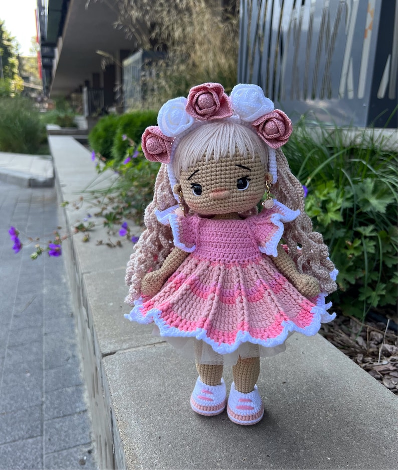 May include: A crocheted doll with pink and white clothing, a pink and white flower crown, and brown hair. The doll is wearing white sneakers and is standing on a gray surface.