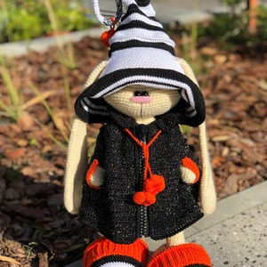 Puede incluir: Un conejito de crochet con un sombrero a rayas blanco y negro, una chaqueta negra con pompones naranjas y botines a rayas naranja y blanco.