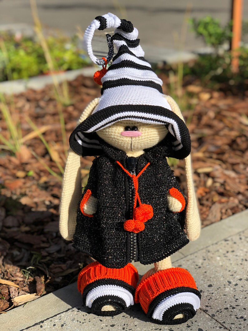 Puede incluir: Un conejito de crochet con un sombrero a rayas blanco y negro, una chaqueta negra con pompones naranjas y botines a rayas naranja y blanco.