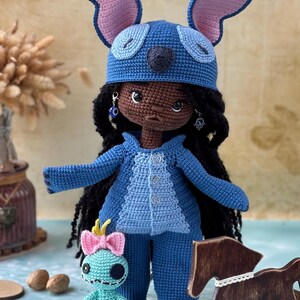 Pu&ograve; includere: Bambola all'uncinetto fatta a mano vestita con un costume blu con grandi orecchie, che ricorda un personaggio dei cartoni animati. La bambola ha i capelli neri e orecchini. &Egrave; presente anche una bambola turchese pi&ugrave; piccola con un fiocco rosa.