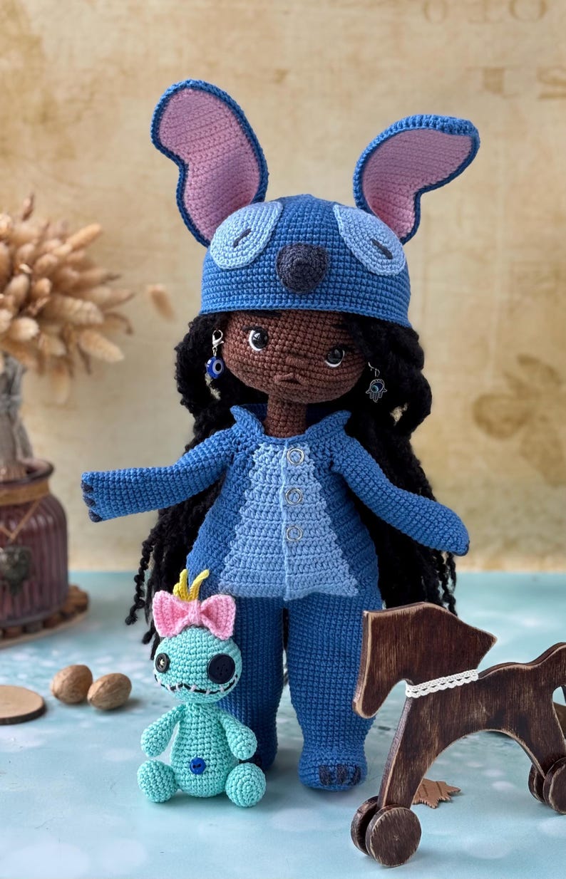 Pu&ograve; includere: Bambola all'uncinetto fatta a mano vestita con un costume blu con grandi orecchie, che ricorda un personaggio dei cartoni animati. La bambola ha i capelli neri e orecchini. &Egrave; presente anche una bambola turchese pi&ugrave; piccola con un fiocco rosa.