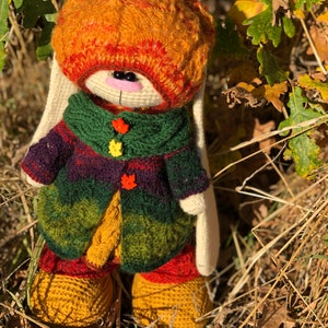 K&ouml;nnte beinhalten: Ein geh&auml;keltes H&auml;schen, das einen mehrfarbigen gestreiften Pullover, eine braune und orangefarbene M&uuml;tze und wei&szlig;e F&auml;ustlinge tr&auml;gt. Das H&auml;schen steht in einem Bett aus trockenem Gras und Bl&auml;ttern.