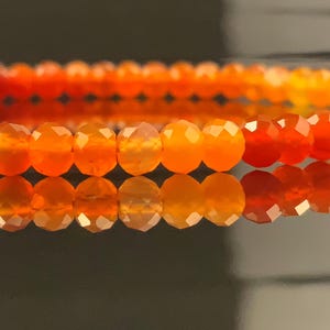 May include: Close-up of a bracelet with faceted orange beads. The beads are strung together, forming a continuous loop. The bracelet reflects on a shiny surface, enhancing the vibrant orange colour. The beads are approximately 0.5 cm in diameter.