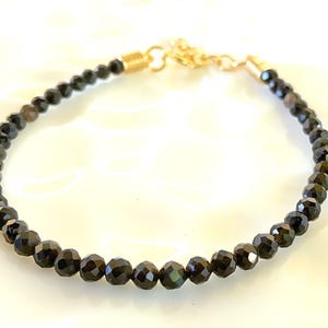 Pode incluir: Um bracelete de pedras preciosas pretas com um fecho dourado. O bracelete é feito de pequenas contas facetadas.