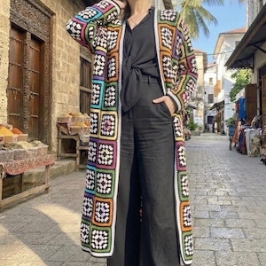 Può includere: Cardigan lungo all'uncinetto colorato con un design patchwork. Il cardigan presenta quadrati di vari colori, tra cui rosa, verde, arancione e viola. La persona indossa pantaloni neri e un top nero. Lo sfondo è una strada acciottolata.