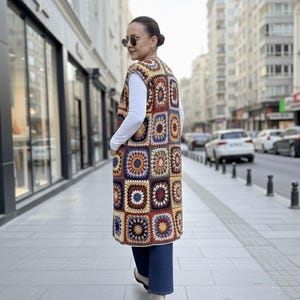 May include: A long, sleeveless crochet vest featuring a patchwork design with squares in shades of brown, blue, yellow, and orange. The vest is worn over a white long-sleeved shirt and blue jeans. The person is walking on a city sidewalk.