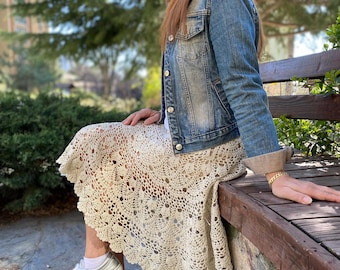 May include: A blue denim jacket with silver buttons worn over a cream-colored, knee-length crochet skirt featuring an intricate floral pattern. The outfit is paired with metallic silver sneakers with white laces and a gold-toned chain bracelet.