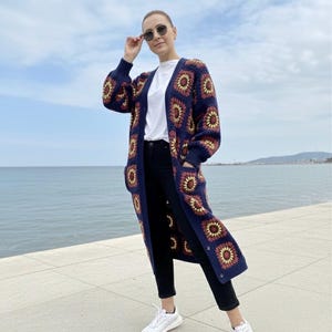 May include: A long, navy blue crochet cardigan with a floral pattern in shades of red, yellow, and brown. The cardigan is worn over a white t-shirt and black pants. The person is wearing white sneakers and sunglasses.