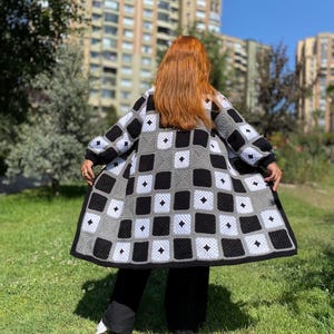 May include: A long-sleeved crochet cardigan featuring a repeating checkerboard pattern in black, white, and gray. The garment is constructed from individual square motifs, each containing a small diamond-shaped cutout, and includes black ribbed cuffs.