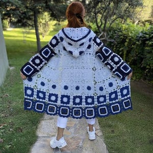 Puede incluir: Un cárdigan de manga larga con capucha hecho de cuadrados de ganchillo granny en blanco y azul marino. La prenda presenta un patrón floral repetitivo en el centro de cada cuadrado, con un borde azul marino sólido a lo largo del dobladillo y los puños.
