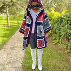 May include: A hooded cardigan made of crocheted granny squares in navy blue, red, white, and gray. The garment features a pointed hood, wide sleeves, and an open front design, layered over a white v-neck shirt, gray jeans, and white boots.