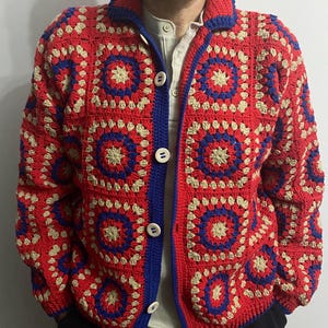 Può includere: Un cardigan all'uncinetto rosso, blu e beige con chiusura a bottoni. Il cardigan presenta un motivo geometrico con colletto e finiture blu. I bottoni sono bianchi. Il capo è di peso medio.