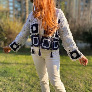 May include: A white crochet jacket with navy blue square accents and tassels. The jacket features a floral pattern on the sleeves and a square pattern on the front. The person is wearing white pants and sneakers.