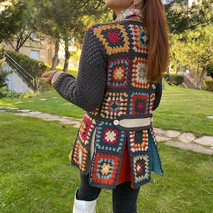 Pode incluir: Um casaco de croché de manga comprida com um padrão de quadrados da avó multicolorido em tons de cinzento escuro, laranja, amarelo e azul. A peça inclui um detalhe de abertura nas costas com dois botões brancos numa faixa horizontal.