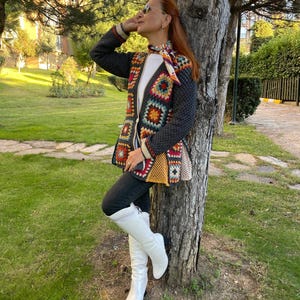 Puede incluir: Un cárdigan de ganchillo de manga larga con base gris oscuro y parches de cuadrados de la abuela en rojo, amarillo, azul y blanco. La prenda incluye una camiseta blanca, pantalones negros, un pañuelo estampado y botas blancas altas.