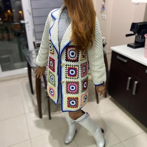 May include: A long-sleeved crochet cardigan made of cream-colored yarn with a granny square pattern featuring colorful geometric motifs in red, yellow, blue, and green. The garment has a lapel collar, blue trim, and two round buttons on the front.