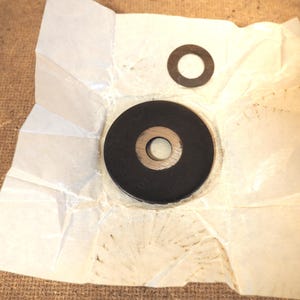 May include: Two black circular washers with a central hole, one large and one small, are displayed on a crumpled piece of white paper. The paper rests on a brown, textured surface.