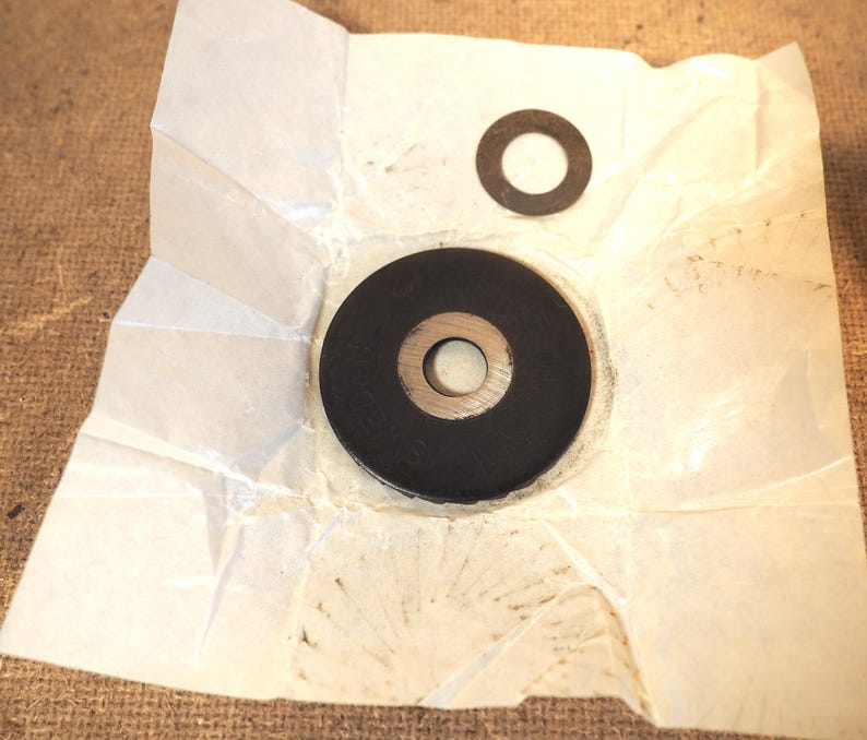 May include: Two black circular washers with a central hole, one large and one small, are displayed on a crumpled piece of white paper. The paper rests on a brown, textured surface.