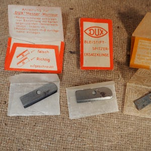 May include: A collection of vintage DUX pencil sharpener replacement blades and packaging. The packaging is orange and white, with German text. The blades are small, rectangular, and metallic. The image shows the blades in their original paper packaging.