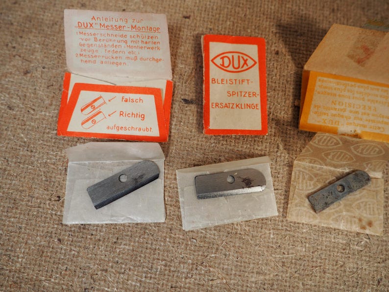 May include: A collection of vintage DUX pencil sharpener replacement blades and packaging. The packaging is orange and white, with German text. The blades are small, rectangular, and metallic. The image shows the blades in their original paper packaging.