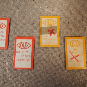 May include: Four vintage DUX pencil sharpener replacement blade cards. Two are red and white, and two are yellow and white. The cards have text in German and French.